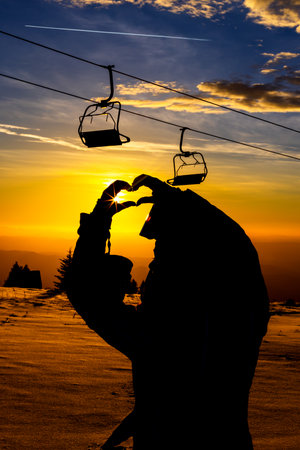 Silhouette of person making heart shape with hands at sunset near ski liftの写真素材