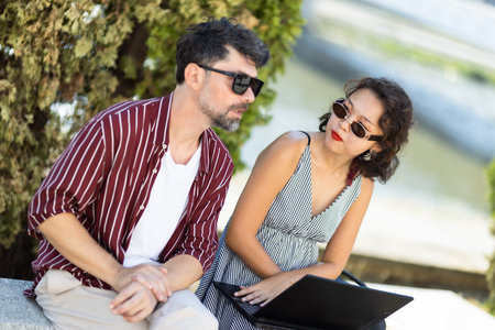 Candid moment of two friends discussing outdoors with sunglasses and laptop during a sunny dayの写真素材
