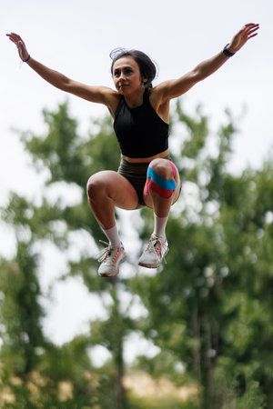 Woman Jumping Outdoors During Physical Activity Wearing Sporty Attire and Knee Strapsの写真素材