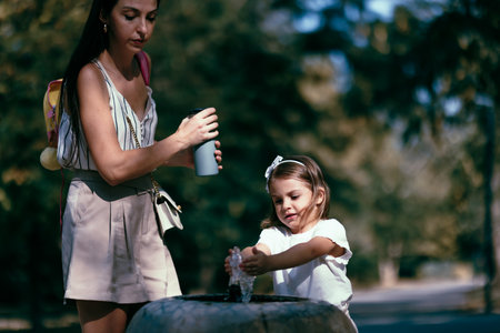 Mother guiding daughter as she plays with water at a park fountain in a sunny outdoor settingの写真素材