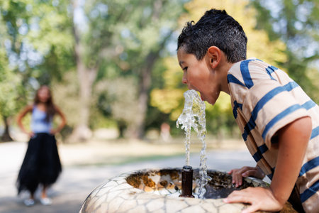 Young boy drinking water from an outdoor fountain during a sunny day in a park settingの写真素材