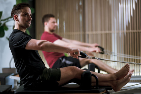 Men engaging in workout routines on reformer pilates machines for strength and flexibility training in a modern fitness studio settingの写真素材