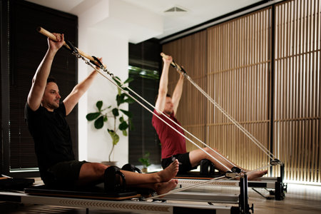 Two men performing Pilates exercises in a modern fitness studio with wooden accents and natural light during a morning sessionの写真素材