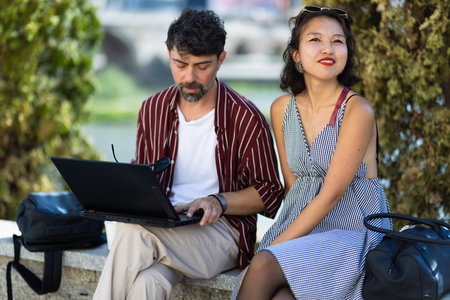 Two people enjoying a sunny day as one works on a laptop and the other relaxes on a bench outdoorsの写真素材