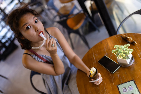 Woman enjoying ice cream at a cafe table with phone and plant, expressive facial expression of surprise and delightの写真素材