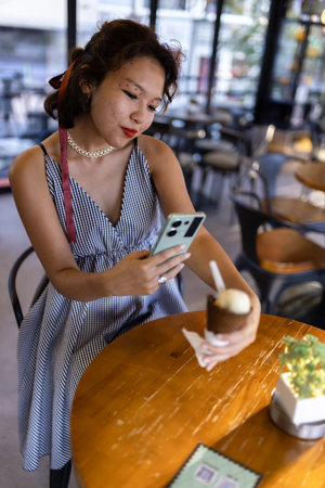 Young woman in a cafe taking a photo of her ice cream with a smartphone, capturing moments and enjoying leisure time indoorsの写真素材