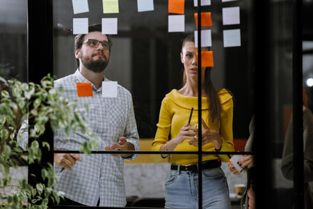 Team brainstorming session in a modern office with sticky notes on the glass wall, showcasing collaboration and creativity during work hoursの写真素材