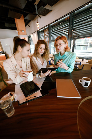 Businesswomen collaborating in a modern cafe, discussing ideas and reviewing documents over coffee in a vibrant urban settingの写真素材