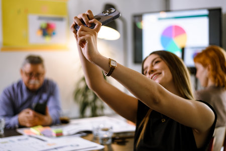 Group of colleagues engaging in a creative meeting while one person takes a selfie with a smartphone in a bright office space filled with colorful charts and documentsの写真素材