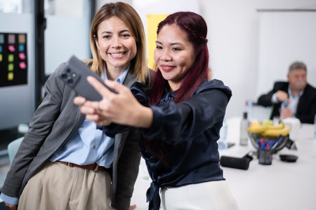 Two professionals taking a selfie together in a modern office workspace, smiling and posing for the camera, showcasing teamwork, camaraderie, and a positive environment among colleagues in a business setting.の写真素材