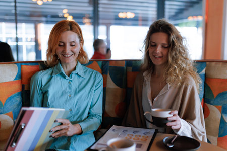 Businesswomen engage in conversation over coffee at a modern cafe while reviewing documents and enjoying each others company in a vibrant settingの写真素材