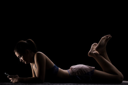 Silhouette of a young woman lying on the floor, absorbed in her smartphone at homeの写真素材