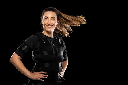 Woman in EMS fitness suit posing confidently for a workout photo against a black background, showcasing strength, fitness, and modern training technologyの写真素材