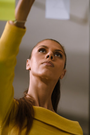 Woman in a yellow sweater engages in creative planning by placing sticky notes on a wall in a bright workspace while concentrating on her ideasの写真素材