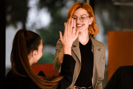 Two women smiling and greeting each other with a high five in a modern office setting during daytimeの写真素材