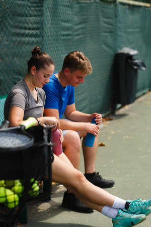 Young athletes resting on a tennis court, cool drinks in hand, enjoying a break during practice in warm weather outsideの写真素材