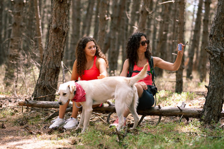 Two women enjoying a break in the forest with a dog on a sunny day, capturing memories on a...