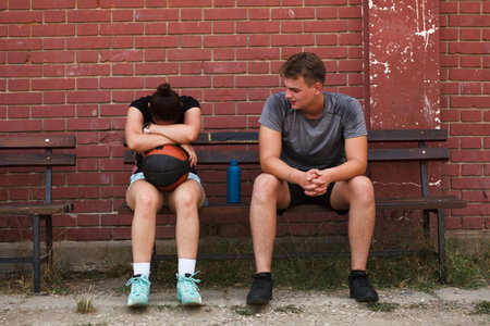 Teen athletes sitting on bench in outdoor basketball court after practice, contemplating the game and future dreamsの写真素材