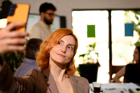 Woman taking a selfie in a bright workspace with colleagues engaged in discussions during a creative...