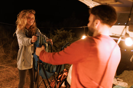 Couple enjoys a nighttime camping experience while setting up chairs under string lights in a serene outdoor settingの写真素材
