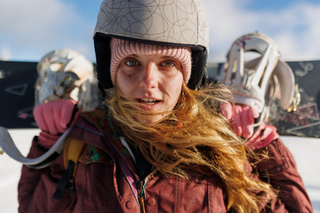 Girl carries snowboard on shoulders in snowy landscape during daylight at ski resort, preparing for...