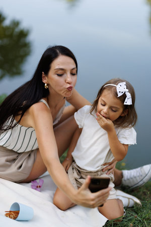 Mother and daughter taking a playful selfie by a serene lake on a warm day