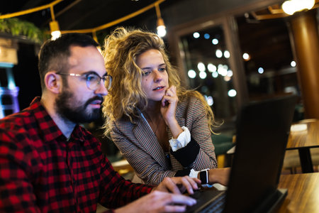Focused colleagues collaborating on a laptop in a cozy, dimly lit coffeeshop. Young man in checkered shirt and woman in blazer intensely engaged in teamwork and discussion, surrounded by soft lighting and reflections.の写真素材