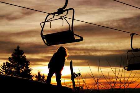 Child stands on a ski lift with sunset in the background while holding a snowboard in an outdoor area during evening hoursの写真素材