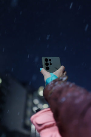 Person holds smartphone under rain at night in urban area with blurred lights in background while...