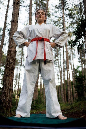 Young girl in karate gi with red belt stands confidently in a forest practicing martial arts and self-discipline barefoot