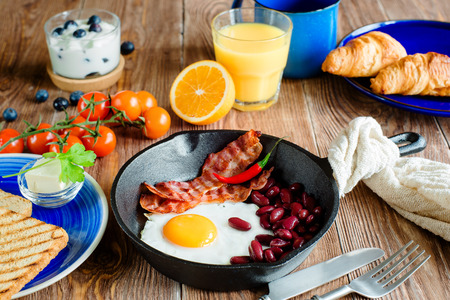 english breakfast in rustic style. fried eggs with beans and bacon on a frying pan. toasts and croissants on a blue plates.の写真素材