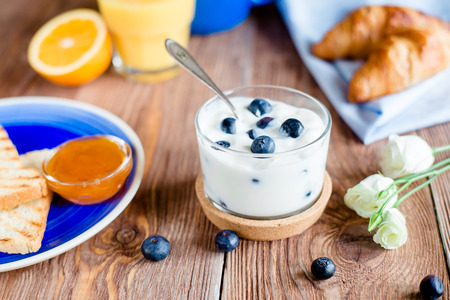 Breakfast with yoghurt with blueberry, toasts with jam and croissants on a blue plate served on wooden tableの写真素材