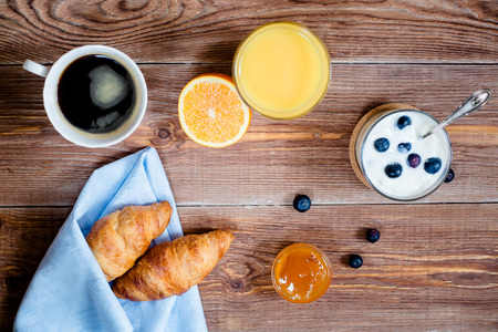 Wooden backgrond with breakfast. cup of coffee, croissants on a blue napkin, yogurt with blueberry, orange juice. top viewの写真素材