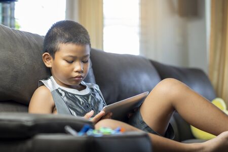 Asian boy playing game or watching cartoons on digital taplet or smartphone on sofa living room.の写真素材