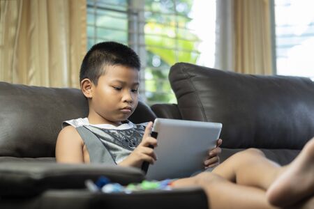 Asian boy playing game or watching cartoons on digital taplet or smartphone on sofa living room.の写真素材