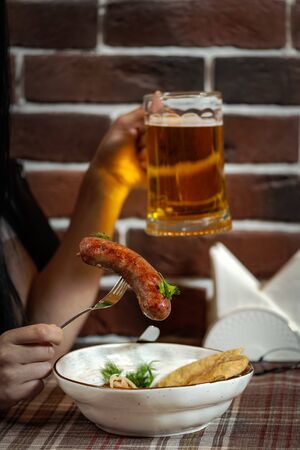 A girl in a pub in one hand holds a mug with beer, in the other a fork with grilled sausagesの写真素材