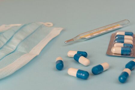 White and blue pills, glass mercury thermometer, face mask on a blue background