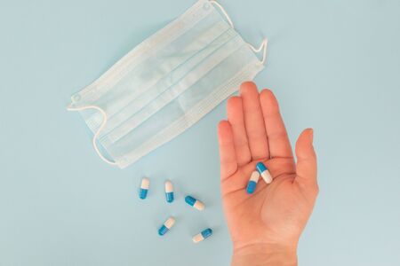 Hands holding white and blue pills, face mask, blue background. Top view