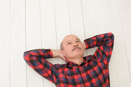 elderly man with a mustache in a plaid shirt lies on a white wooden floor and dreaming, or planning somethingの写真素材