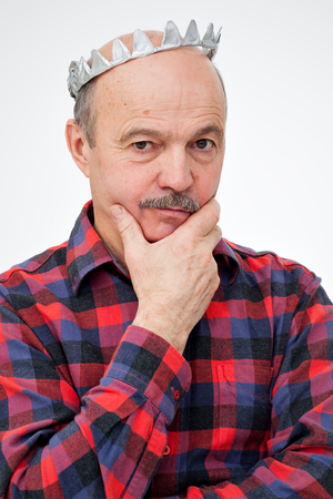 Elderly man with a paper toy crown stands with folded hands on his head. Thoughtful look forward.の写真素材