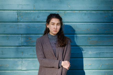 A beautiful Asian girl with long hair and a warm sweater is standing by the wooden blue wall. She looks forward mysteriouslyの写真素材