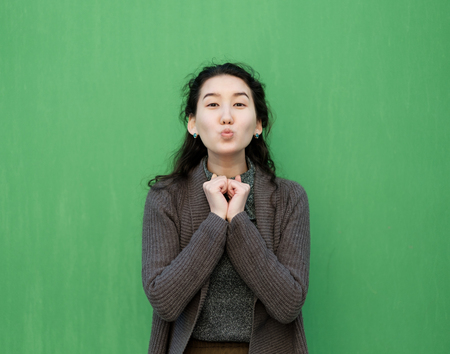 Beautiful Asian girl sends an air kiss. She is standing in a warm sweater near the green wallの写真素材