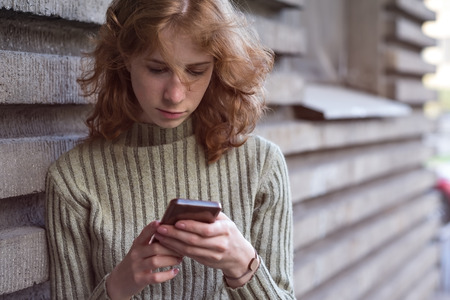 A red-haired girl with curls stands on the street with her phone and looking for the right information in internetの写真素材