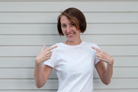 A beautiful girl with freckles in a white T-shirt is standing by the wooden wall. She points to herself with her fingers, clarifying the choiceの写真素材