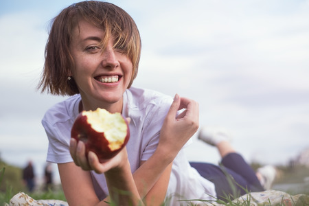 Very beautiful caucasian model eating red apple in the park. Outdoors portrait of pretty young girl lying on green grassの写真素材