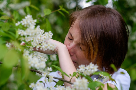 A beautiful girl stands by the blossoming bird cherry and sneezes. She suffers from an allergy to flowering in the spring.の写真素材