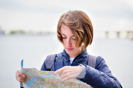 A beautiful girl in a denim jacket is holding a map and trying to understand where she is. Lost in an unfamiliar city. Making a route for the excursion.の写真素材