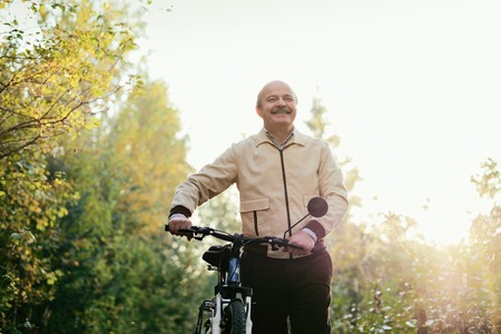 Senior caucasian man go for a walk with bike in countryside. Green trees on background. He is happy and active.の写真素材