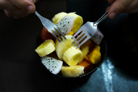 Bowl of healthy fresh fruit salad on wooden background. Man eating it with two forks. Dark color imageの写真素材