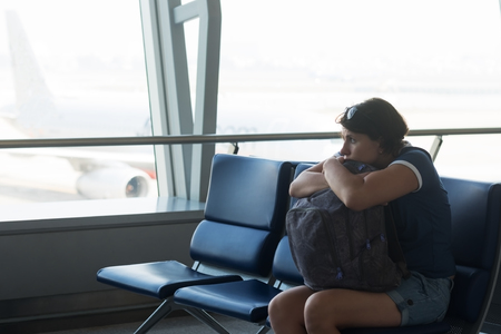 Tired caucasian woman in airport hall. Girl waiting her flight at airport terminal, holding her bag in hands, sitting on chair and looking on the window. She is afraid of airplaneの写真素材